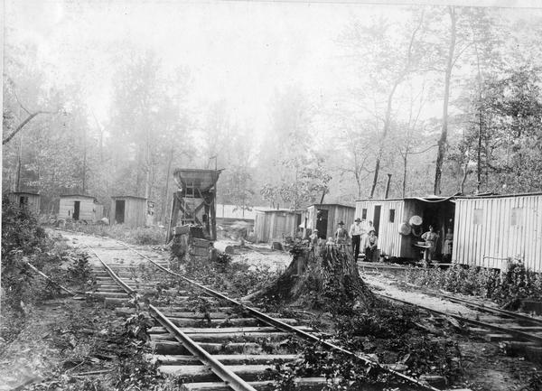 Housing for employees of logging and lumber operations associated with International Harvester. The housing consists of small one-room shacks in a wooded area along railroad tracks. The workers' and their families are also present.