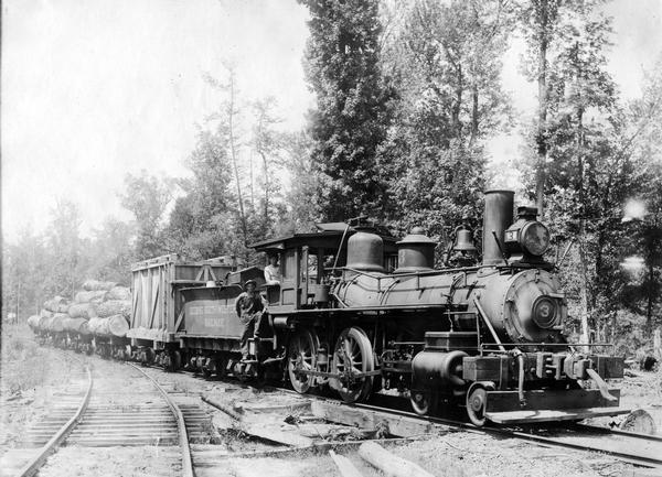 Two workers perched on a steam locomotive pulling railroad cars loaded with logs. The coal car bears the name "Deering Southwestern Railway." The railway was originally used for logging operations associated with the Deering Harvester Company. The logging operations became part of International Harvester after 1902.