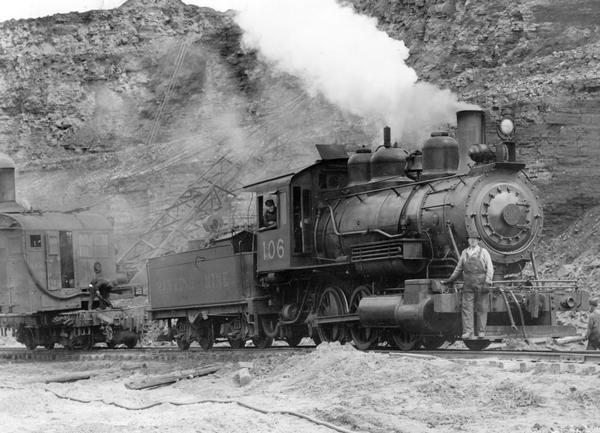 Steam locomotive moving an overhead crane used to lay rails for International Harvester's Hawkins Mine. One man is standing on the right, and another man is looking out the locomotive window.
