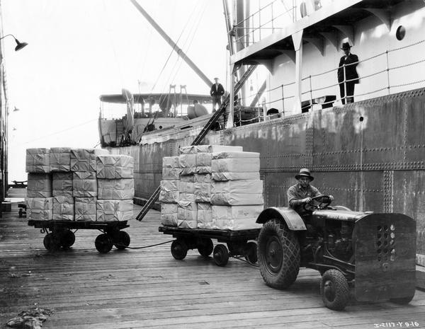 Dock worker hauling two small trailers of cargo with a McCormick-Deering I-12 industrial tractor next to a docked ship. The tractor was owned and operated by the Port Authority.