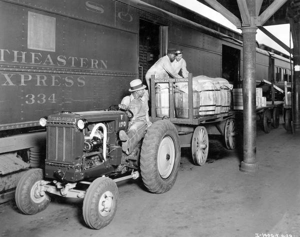 African American workers load barrels of freight  onto a Southeastern Express rail car at a Jacksonville rail station. One worker is operating an International I-12 industrial tractor while the other workers remove freight  from the attached wagon. The tractor was owned by the Southeastern Express Company.