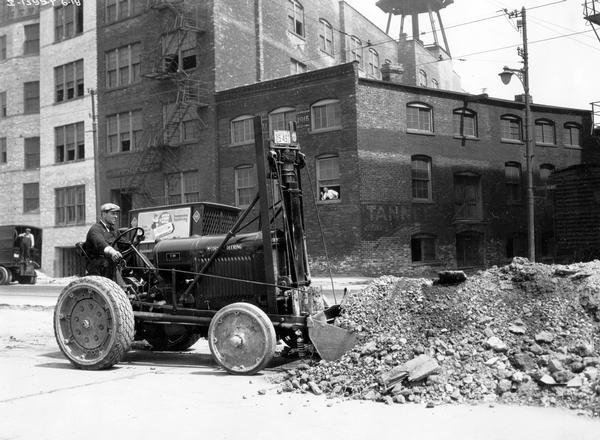 Commonwealth Edison worker using a McCormick-Deering I-30 industrial tractor with a loader to scoop gravel off of a city street. The loader was manufactured by the Frank G. Hough Company.