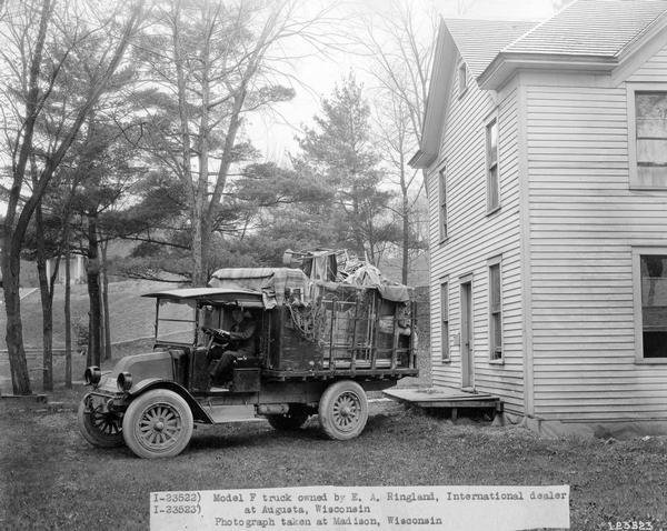 Man behind the wheel of an International model F truck loaded with furniture in front of a residential home. The truck was owned by E.A. Ringland, International dealer at Augusta, Wisconsin.