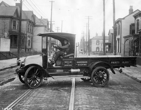 Man in the driver's seat of an International Model F or 31 truck parked in the middle of a city street. The truck was owned by Caldabaugh Bros., proprietors of hardware, roofing, paints and glass materials.