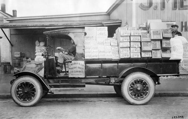 Two men hauling boxes of grocery items in an International Model K-41 truck. Items in the back of the truck include Jell-O, Bon Ami cake, Warner's spaghetti and macaroni, Ideal raisins, and Queen Esther flour. The truck was owned and operated by J. Meunier and Son, wholesale grocers.