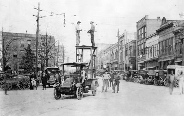 Two men working on trolley car electrical wires (catenary) in the middle of a street. The men are standing on a platform in the back of an International Model H  truck as other people in the street are looking on. The Model H eventually became the Model 21.