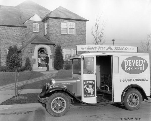 Milkman delivering a bottle of milk to a woman at her residential home. Parked in the foreground along a curb is an International Model M-2 milk truck owned by Pevely Selected Milk Co. An advertisement for "Baby First Selected Milk" is painted on the side of the truck. The M-2 was designed exclusively as a milk delivery vehicle. It had a 118 inch wheelbase, 186 cubic inch four cylinder engine, Warner T-9 transmission, and IHC rear axle. The body was built with a special treatment of its wood components to resist lactic acid damage.