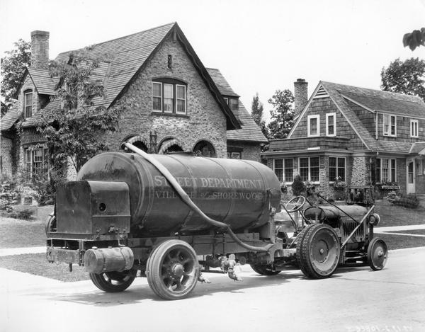 McCormick-Deering 10-20 industrial tractor parked along a curb in a residential neighborhood. The tractor is attached to a trailer with a tank that bears the words "Street Department, Village of Shorewood."