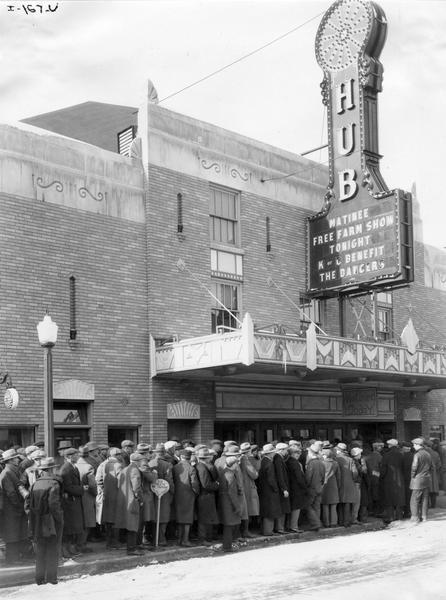 Large crowd lined up outside the Hub theatre for a "free farm show" matinee, most likely "Romance of the Reaper". The movie was produced by International Harvester to celebrate the "reaper centennial" (the anniversary of the invention of the first mechanical reaper by Cyrus McCormick).