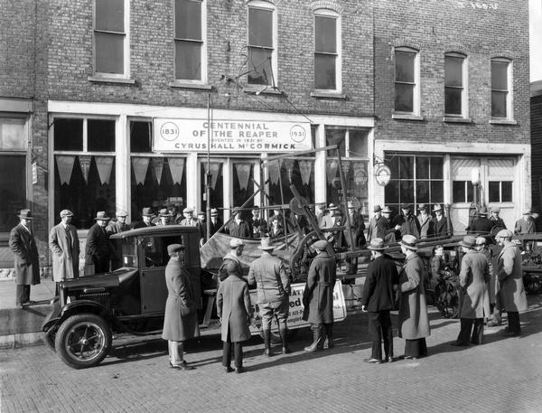 Crowd examining a full size reproduction of the "first reaper" displayed on the bed of an International "Six Speed Special" truck parked in front of an International Harvester dealership. The company created the reproductions and displayed them around the United States for the "reaper centennial" celebration (the anniversary of the invention of the mechanical reaper by Cyrus McCormick).