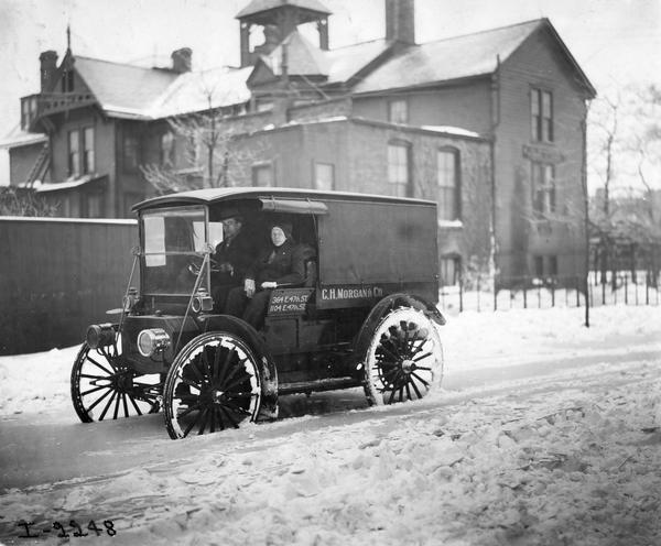 Two young men are driving an International Auto Wagon through a snow-bound residential street. The truck was owned by C.H. Morgan & Co.