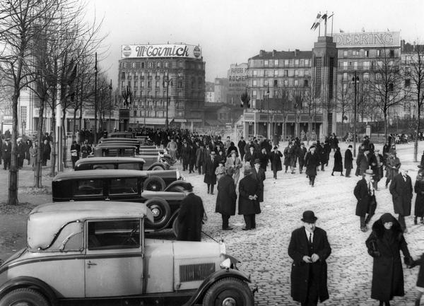 Elevated view of a crowd of people streaming from the direction of two large buildings labeled "McCormick" and "Deering" in what appears to be a French city.