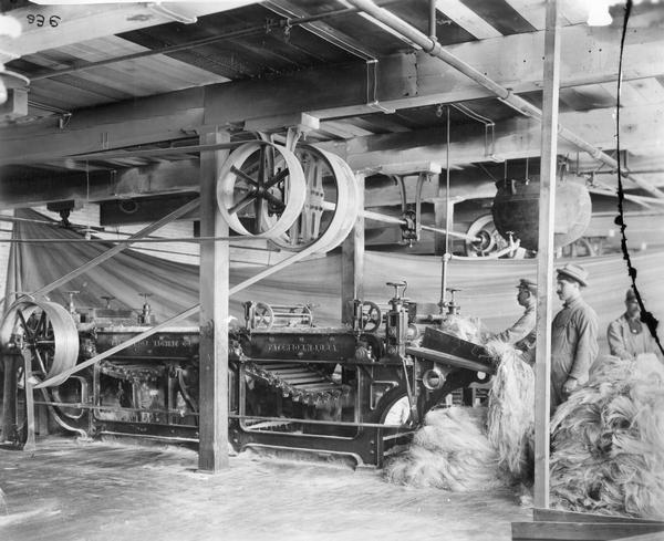 Workers using a large machine to comb twine fibre (fiber) - the first process preparatory to spinning - at International Harvester's Osborne Twine Mill. The Osborne Works was owned by D.M. Osborne Company until 1903, when it was purchased by International Harvester. The factory was later known as "Auburn Works."