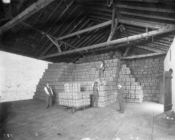 Three workers and a supervisor stacking bundles of twine all the way to the roof in a warehouse at International Harvester's Osborne Twine Mill. The Osborne Works was owned by D.M. Osborne Company until 1903, when it was purchased by International Harvester. The factory was later known as "Auburn Works."