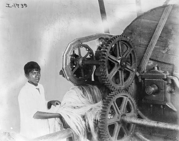 Boy removing a bundle of twine from a decorticator machine at an International Harvester factory in the Yucatan, Mexico. Accompanying text reads: "No guard. Do not use."