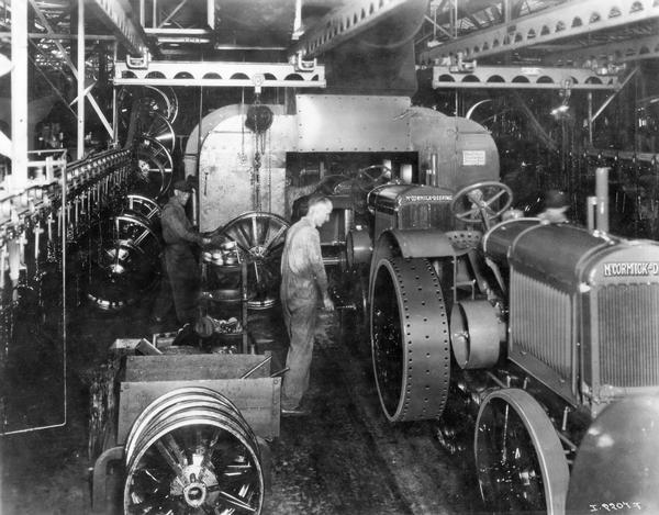 Factory workers installing front wheels on McCormick-Deering 10-20 tractors as they come through a painting booth on an assembly line at International Harvester's Tractor Works. The wheels arrive by overhead conveyor.