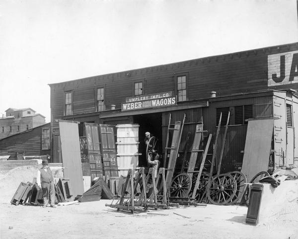 Two men unloading Weber wagon parts from a railroad car in front of the Umpleby Implement Company while a portly man in a bowler hat is gesturing from a distance. Umpleby was a Weber Wagon dealership. Note on back of original print states: "sent in by Springfield, Ill. for use on card to dealer, 7-1-16."
