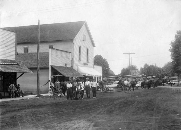 Farmers in the street of a rural town with their International Harvester horse-drawn mowers and binders on "McCormick Day." The event was organized by the Red Front Implement Store to publicize the delivery of new machines to customers. The dealership was owned by G.A. Auble. One man is sitting in a wheelchair.