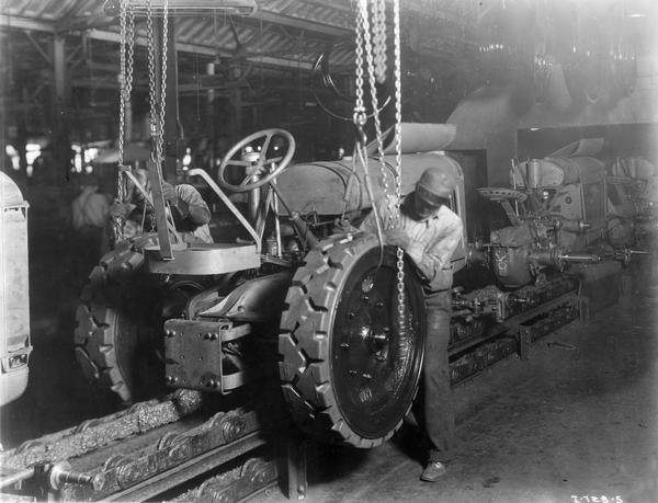 Two factory workers attach the rear wheels to an industrial tractor frame on an assembly line at International Harvester's Tractor Works. Original caption in "Harvester World" magazine reads: "A doughty industrial gets a pair of 1700-pound solid steel wheels as it enters the painting booth on its way over the final 250 feed of the assembly line."