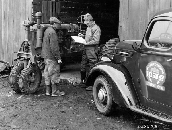 Serviceman Harold Finch filling out an "Inspector Form C-3219" for a Farmall F-20 tractor on the farm of H.B. Ayers as Mr. Ayers looks on. Mr. Finch's International D-series truck, with a "McCormick-Deering Service" emblem on the door, is in the foreground at right. Mr. Finch is wearing the official IH service uniform.