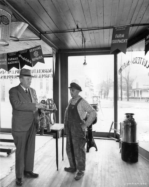 Salesman Claude De Kay demonstrating the functionality of a McCormick-Deering milker to customer H.M. Arnold against the backdrop of the store's plate glass windows. De Kay was co-owner of Prosser and De Kay, Inc., an International Harvester dealership.