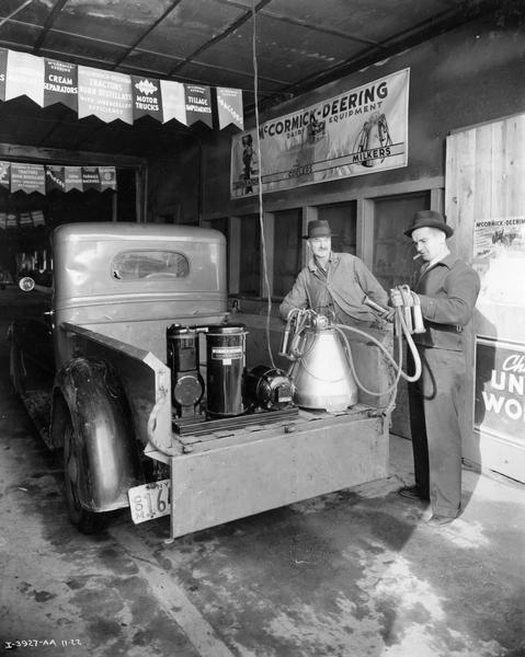 McCormick-Deering dealer Edward Gumienick smoking a cigar as he is loading dairy equipment into his International truck for a sales "canvassing" trip. His serviceman Fred Keith (left) is looking on. Advertising posters and banners are in the background.