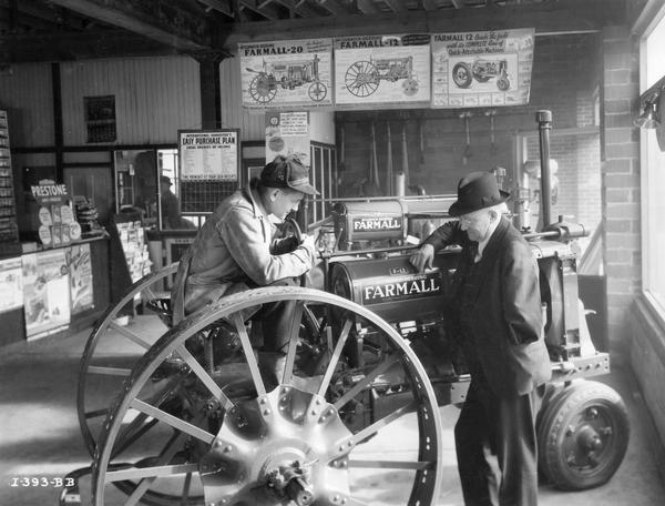Dealer P.W. Stonebrig(?) and his son posing as a dealer explaining a Farmall F-12 tractor to a customer. A Farmall F-20 tractor and several advertising posters are hanging from the ceiling in the background.