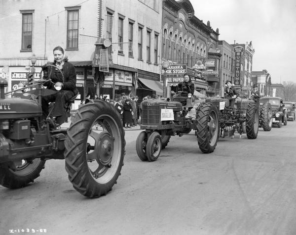 Farmall H and M tractors driven in a parade through a downtown street. The tractors are driven by two women and two men. One of the men has a small child on his lap. There is a crowd behind them on a street corner, and above them is an elaborate sign on the corner of a building in the shape of a windmill.