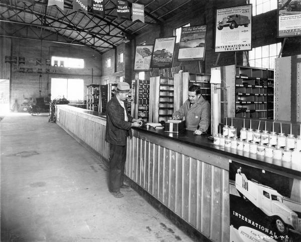 Customer buying parts at the service desk of an International Harvester dealership. Advertisements for International trucks are posted above the counter.