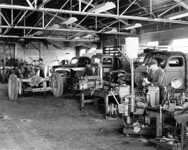 Mechanics at work in the service shop of an International Harvester dealership, working among several disassembled McCormick-Deering tractors, engines, and International trucks.