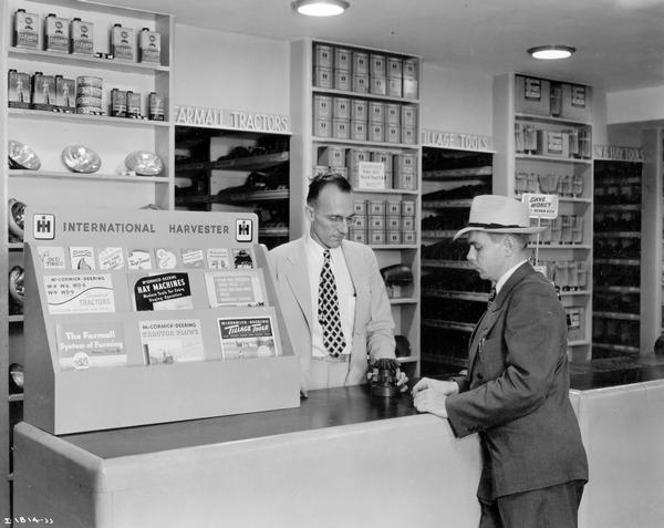 Man standing at the service desk of an International Harvester dealership. A clerk is behind the counter. Advertising brochures are displayed on the left edge of the counter.