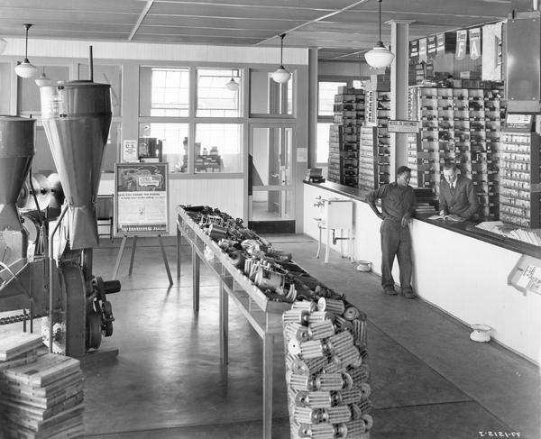 Slightly elevated view of a customer at the service counter of an International Harvester dealership. A bin of oil filters, McCormick-Deering hammer mills, and an advertisement for the U.S. Truck Conservation Corps are displayed.