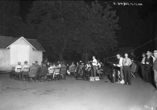 Rural farming crowd gathered to watch an International Harvester film on the side of a barn. The event was staged by J.H. Matthews, an International Harvester advertising man, who used outdoor evening movies at farmers' homes to promote IH products and services.  A man is serving ice cream from a large container at right.