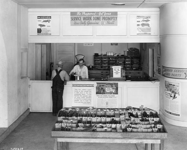 Customer standing at the service counter of an International Harvester dealership. Advertising posters for the U.S. Truck Conservation Corps and WWII scrap metal drives are posted around the counter.