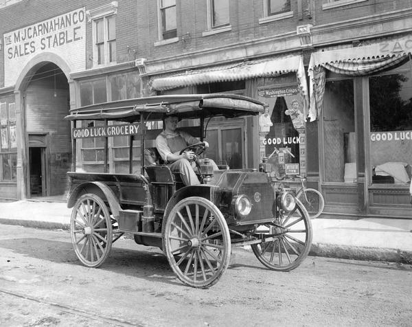 Man sitting behind the wheel of an International Model M grocery truck in front of the Good Luck grocery store. A sign on the back of the truck reads: Good Luck Grocery". The store is next to the M.J. Carnahan Co. sales stable.