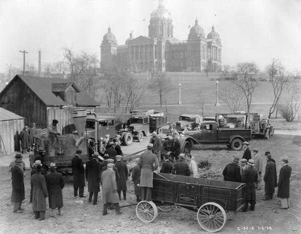 Elevated view of men gathered for a McCormick-Deering farm machine demonstration on grounds across from the Iowa capitol building. Equipment on view includes Weber Wagon, International trucks, McCormick-Deering hammer mill, and McCormick-Deering tractor.