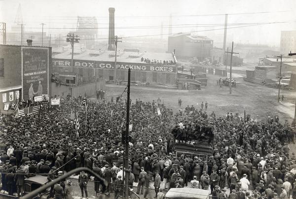 Elevated view of hundreds of people, possibly factory workers, gathered to hear speeches outside International Harvester's Deering(?) Works. The event appears to be related to World War I. Flags adorn the speakers' stage and there appear to be soldiers in the crowd. A sign near the stage reads: "Why not keep saving? Buy victory loan bonds." The Deering Works was built by William Deering for the Deering Harvester Company in 1880 and became International Harvester's Deering Works in 1902. The factory was located at Fullerton and Clybourn Avenues and closed in 1933.