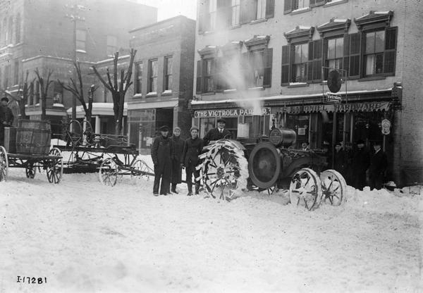 Men gathered around an International Harvester Mogul 10-20 HP tractor on a snow-covered city street. A grader is attached to the tractor to plow snow. The tractor is idling in front of the Samoniel Hardware store and "Victrola Parlor."
