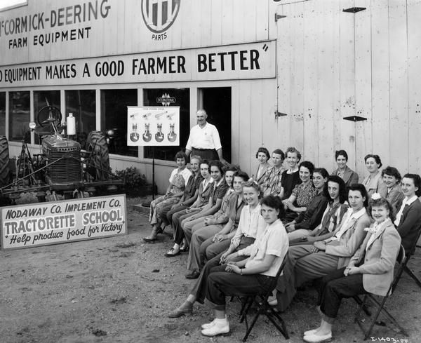 Women assembled for a Tractorette School at Nodaway Co. Implement Co., an International Harvester dealership. Instructor and dealer John Schneider is conducting an outdoor lesson on the four stroke engine. A Farmall tractor is parked outside the storefront. The company announced the "Tractorette" program in 1942. The plan was intended to address the farm labor shortage created by U.S. war mobilization. The plan called for local dealers to offer free training for thousands of farm women and girls in the operation of tractors and other farm machinery.