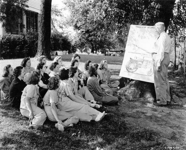 Women sitting on grass watching a lecture on maintenance of Farmall A and Farmall M tractors. The lecture was part of International Harvester's "Tractorette" program. The company announced the "Tractorette" plan in 1942. The plan was intended to address the farm labor shortage created by U.S. war mobilization. The plan called for local dealers to offer free training for thousands of farm women and girls in the operation of tractors and other farm machinery.