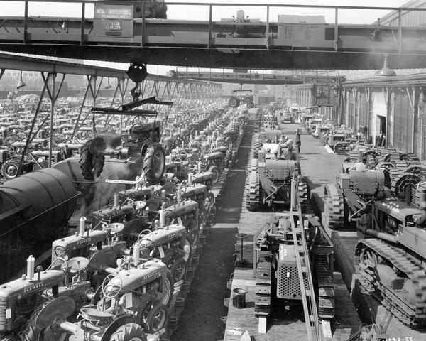 Elevated view of men loading hundreds of new Farmall B, Farmall A and International crawler tractors (TracTracTors) onto railroad cars with a crane outside International Harvester's Tractor Works. Some of the crawler tractors appear to be bound for the U.S. military. The factory was located at 2600 West 31st Boulevard.