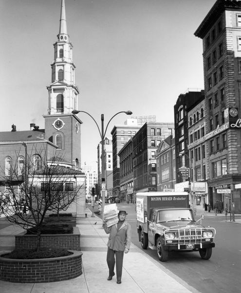 Man delivering a stack of Boston Herald newspapers from an International C-series truck parked along the curb in the downtown Boston area at Boston Common. In the background is the Park Street Church.