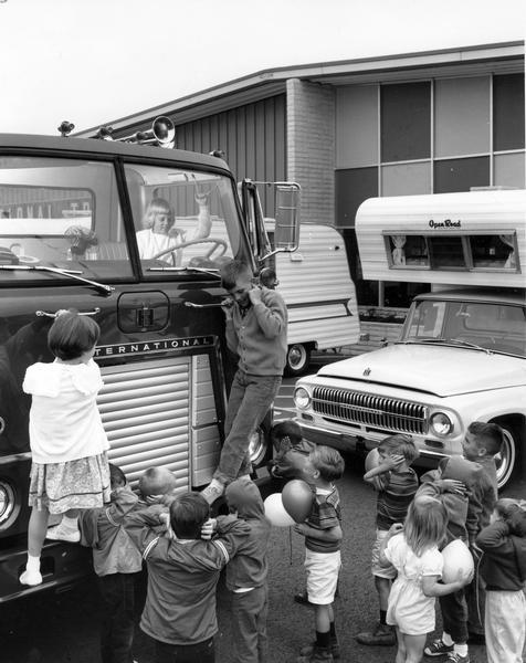 A group of children are covering their ears as a young girl sounds the horn of an International truck. Some of the children are holding balloons in their hands.