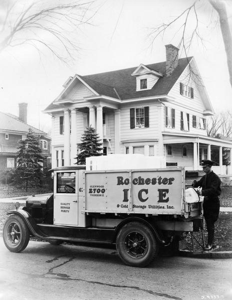 Man removing a block of ice from an International truck for delivery in a residential neighborhood. The house in the background is at 462 Seneca Parkway. The truck was owned by Rochester Ice and Cold Storage Utilities, Inc.