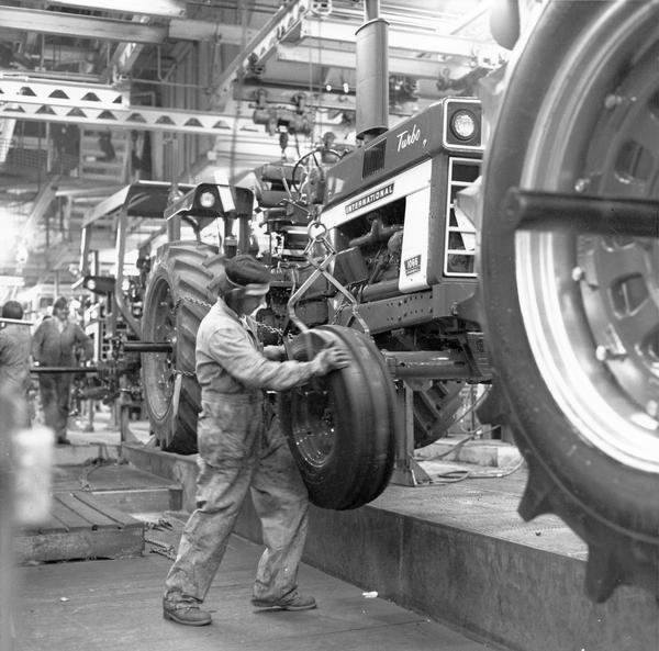 Worker attaching the right front wheel to an International 1066 Turbo tractor along an assembly line.