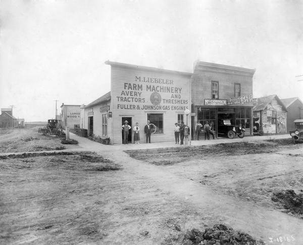 View across unpaved road towards men, boys, and a young woman standing on a sidewalk in front of the storefront of M. Liebeler Farm Machinery and Garage. The buildings advertise Avery tractors and threshers, Fuller & Johnson gas engines, McCormick farm machines, and Dodge cars. The buildings occupy the corner of a dirt street in a rural town.