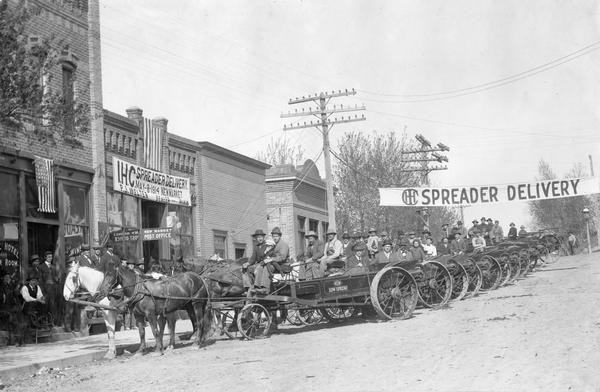 Farmers and their families lined up in the street of a rural town for a "spreader delivery" day organized by International Harvester dealer, P.J. Welter. The families are sitting in their new horse-drawn manure spreaders. The event was organized to publicize the delivery of new machines.