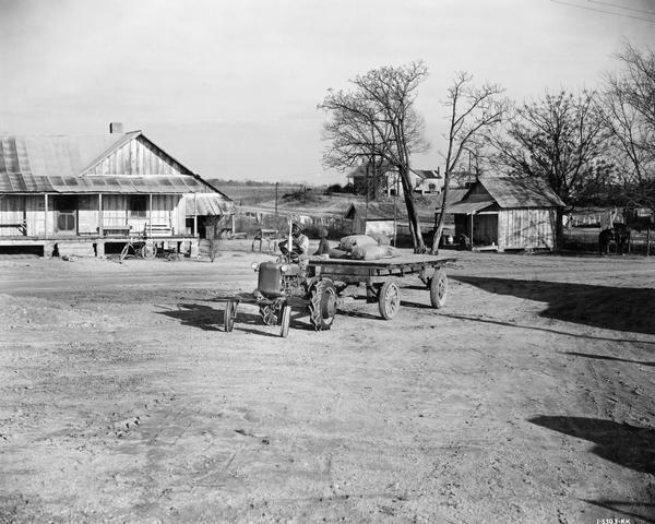 African American farmer pulling a trailer with a Farmall Cub tractor on McQueen Smith Farms. The trailer is loaded with large sacks, and a child is sitting on one edge.