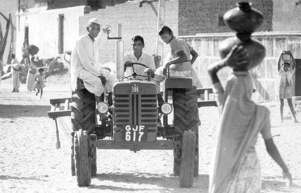 Three men riding an International Harvester tractor as women and children carry jars and baskets along a road in India. The original caption reads: "In 1965, International Harvester Company entered into a joint venture with local interests, and opened a new tractor plant at Kandivli, an industrial area outside of Bombay."