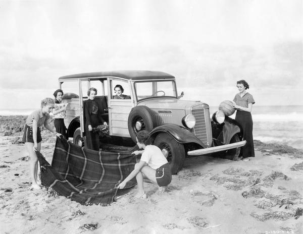 Women preparing for a picnic at the beach with their International C-1 wood-paneled ("woody") station wagon.